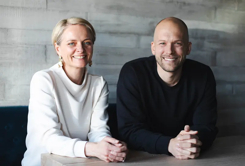 Eine Frau in einem weißen Oberteil und ein Mann in einem schwarzen Sweatshirt sitzen zusammen an einem Holztisch vor einer grauen Betonwand.