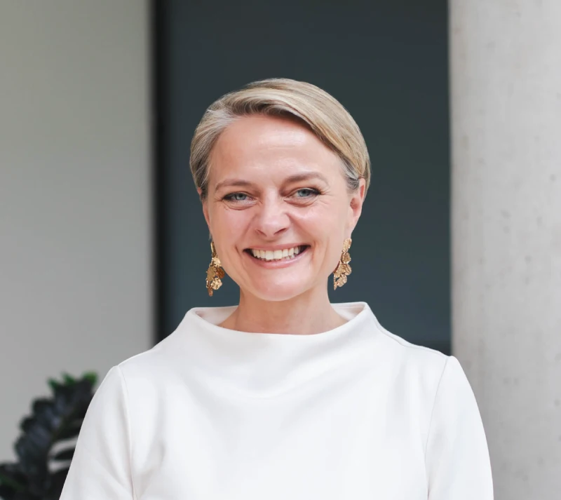 Eine Frau mit kurzen blonden Haaren, die ein weißes hochgeschlossenes Oberteil trägt, steht vor einem modernen Hintergrund mit dezenten Farben.