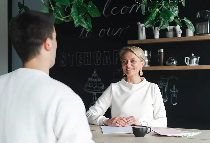 Zwei Personen unterhalten sich an einem Tisch, umgeben von Pflanzen und einer gemütlichen Atmosphäre. Dokumente und eine Kaffeetasse sind vorhanden.